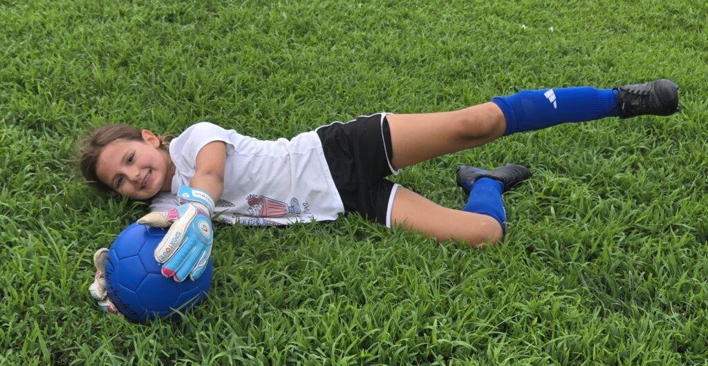 A young soccer goalkeeper diving on the grass to catch the ball.